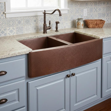 Copper farmhouse sink in a kitchen with gray cabinets and a window above.
