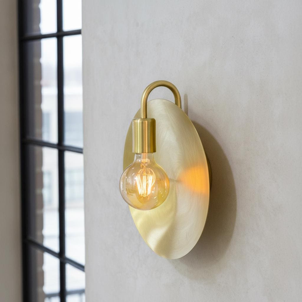 Wall-mounted light fixture with a gold arm and beige shade on a white wall.