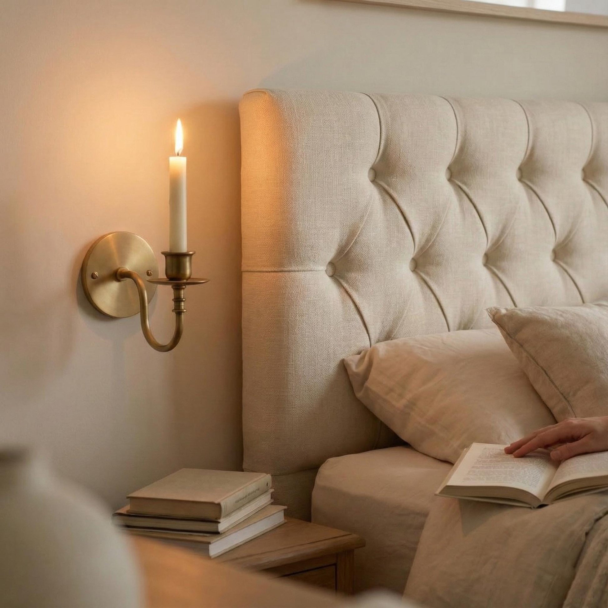 Person reading a book on a bed with a candle and books on a nightstand.