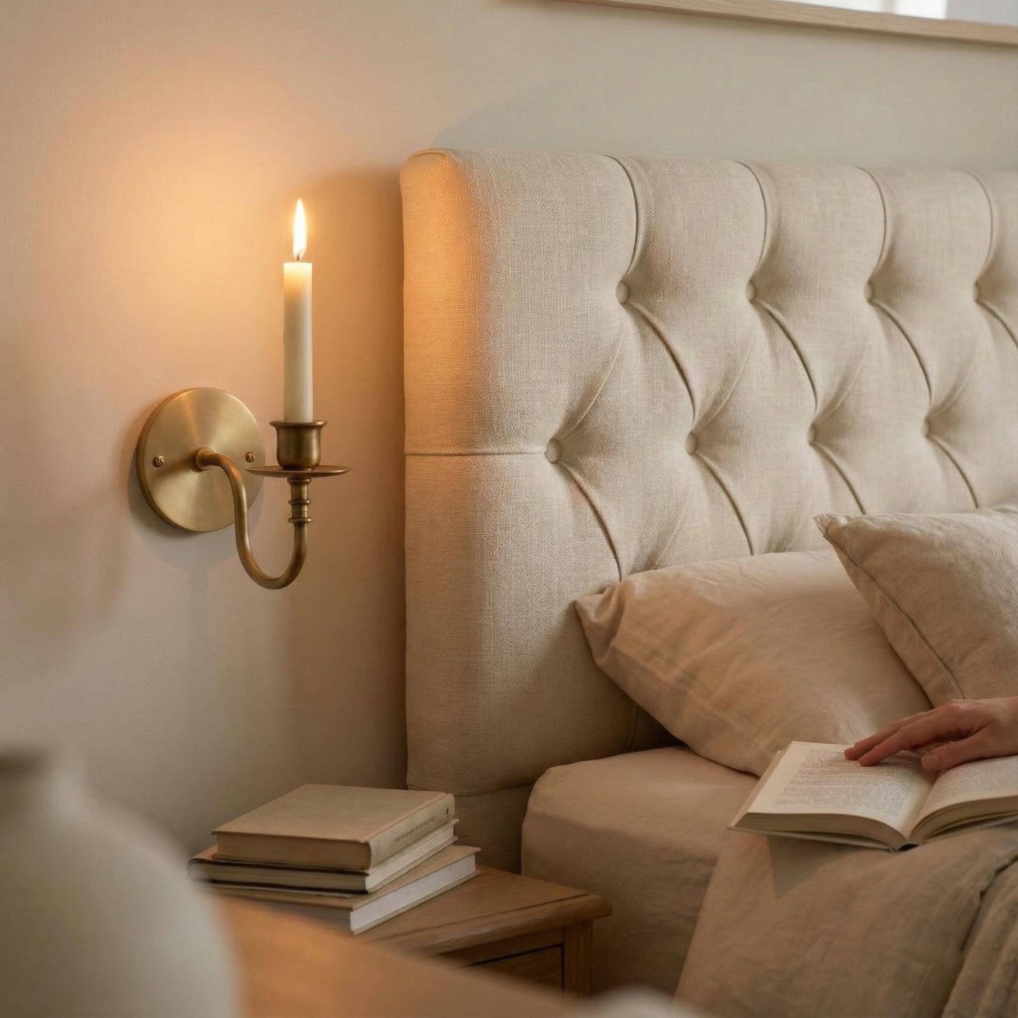 Person reading a book on a bed with a candle and books on a nightstand.