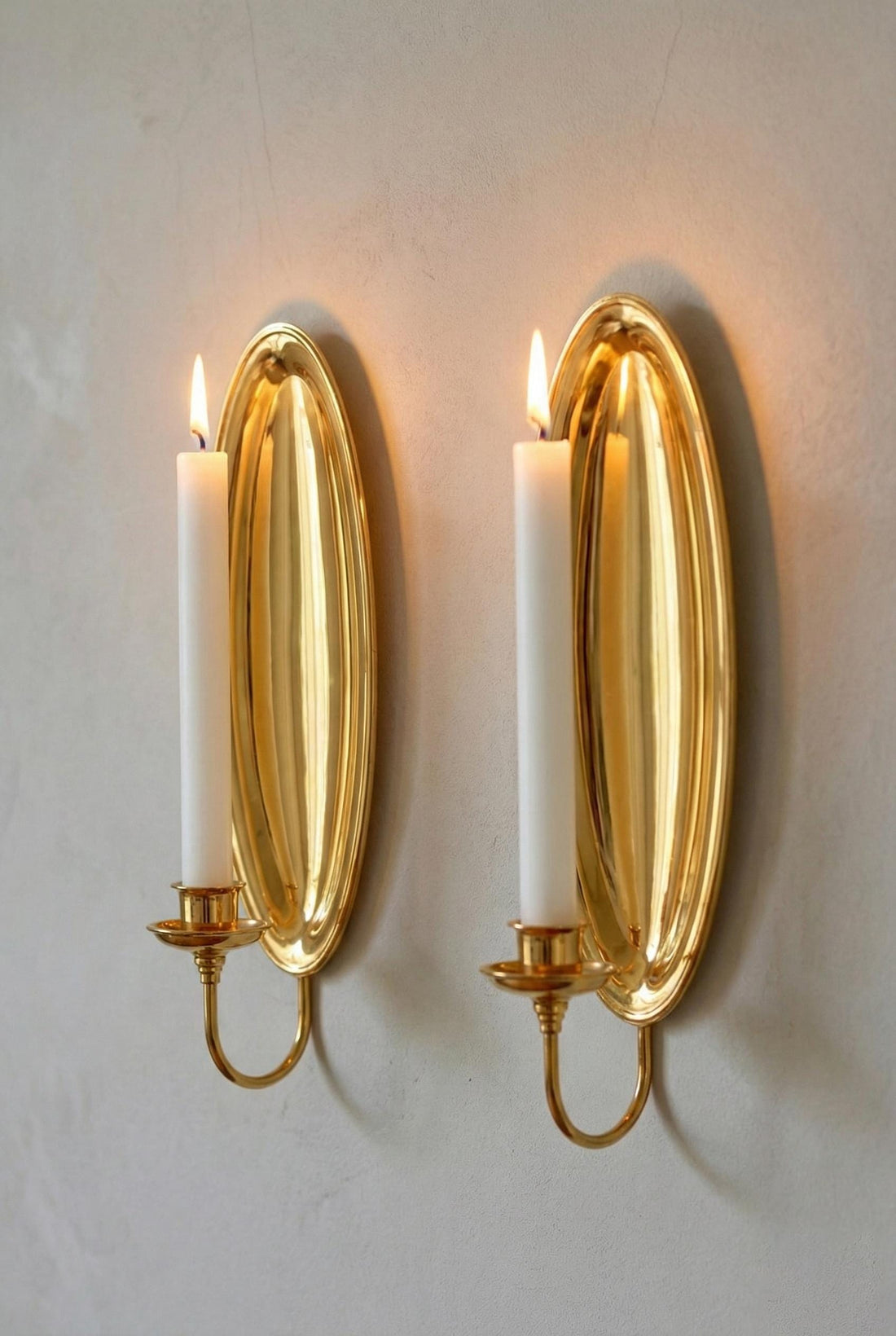 Two gold wall sconces with lit candles on a light gray wall.
