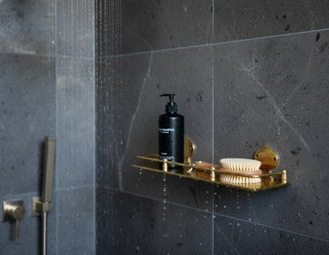 Shower area with a gold soap dish holding a black bottle and a white brush against a dark tiled wall.
