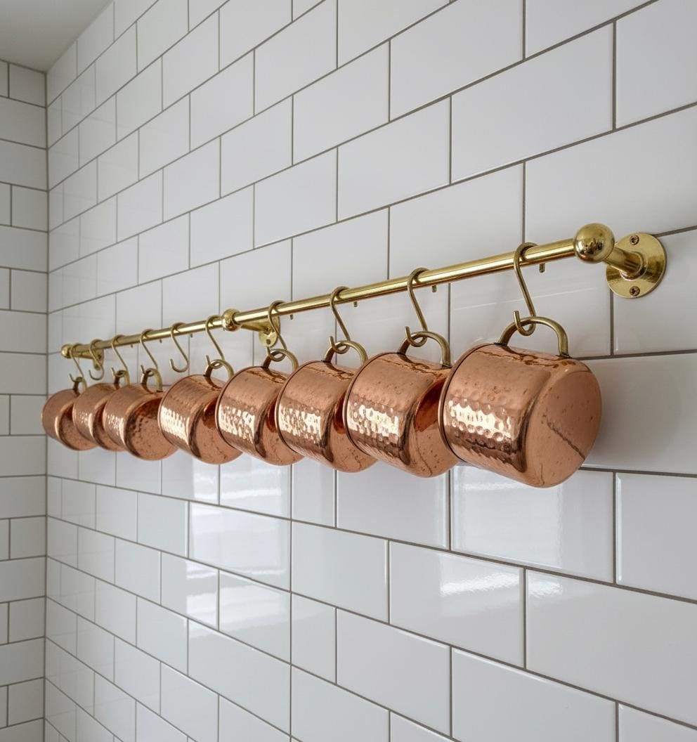 Set of copper measuring cups hanging on a gold rod against a white tiled wall.