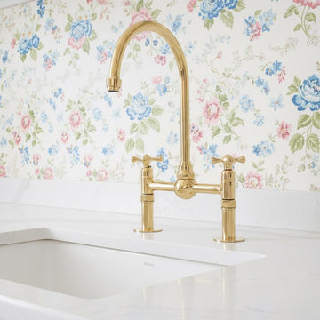 Gold kitchen faucet on a white countertop with floral wallpaper in the background
