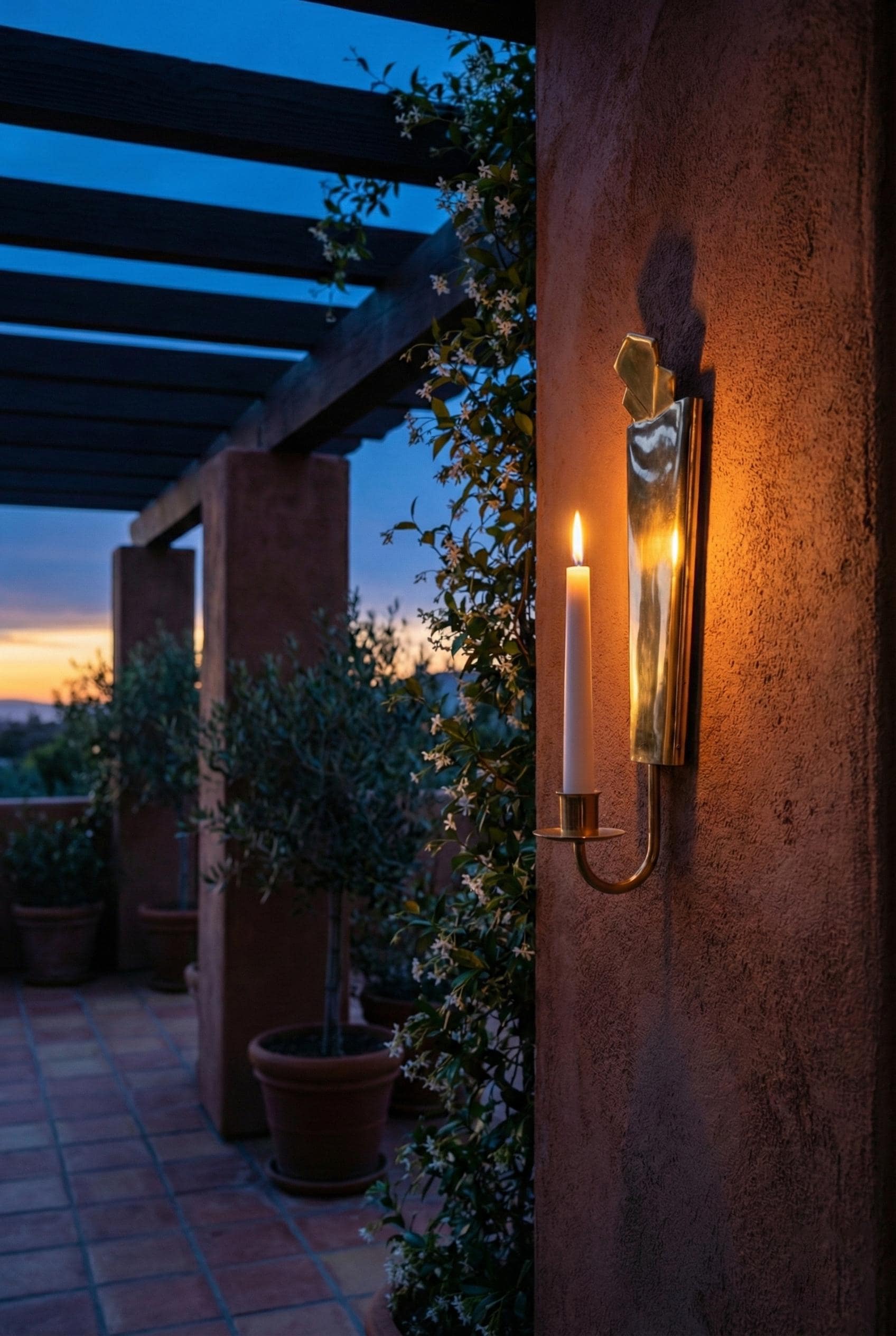 Outdoor setting with a wall-mounted candle lantern on a warm-toned wall, evening sky, and plants.