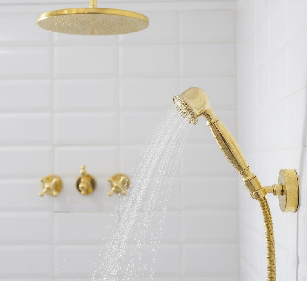 Gold shower head and hose with water flowing against a white tiled wall.