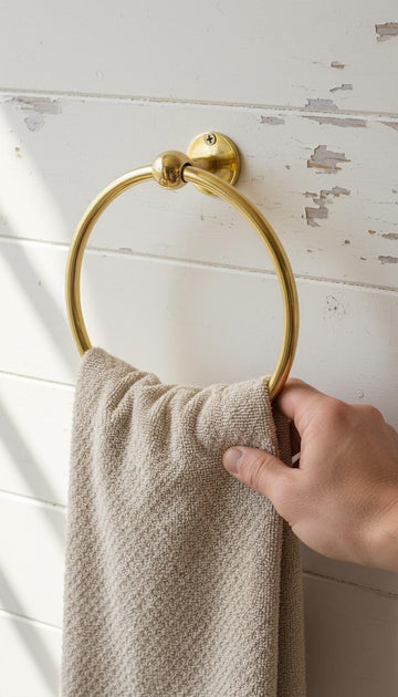 Gold towel ring on a white wooden wall with a beige towel and hand.