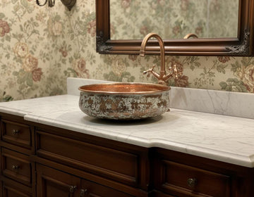 Copper sink on a marble countertop with floral wallpaper and a mirror above.