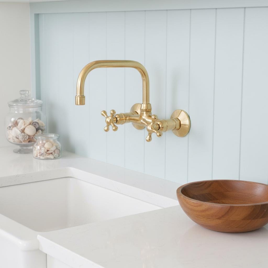 Gold faucet on a white kitchen wall with a wooden bowl and jars below.