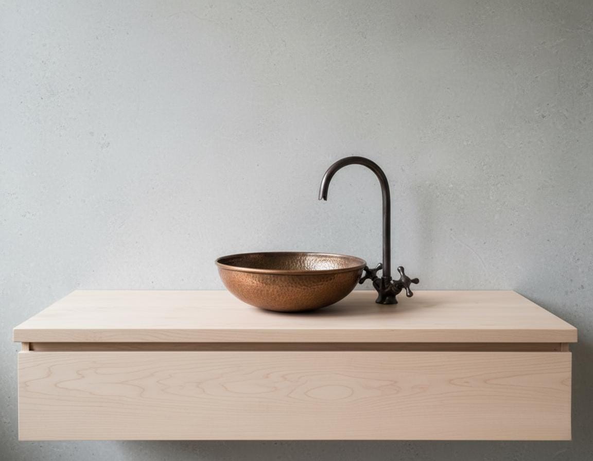 Copper sink with black faucet on a light wood vanity against a gray wall.