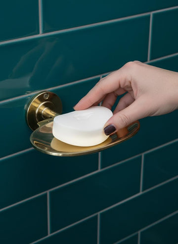 Hand holding a bar of soap over a gold soap dish against a blue tiled wall.