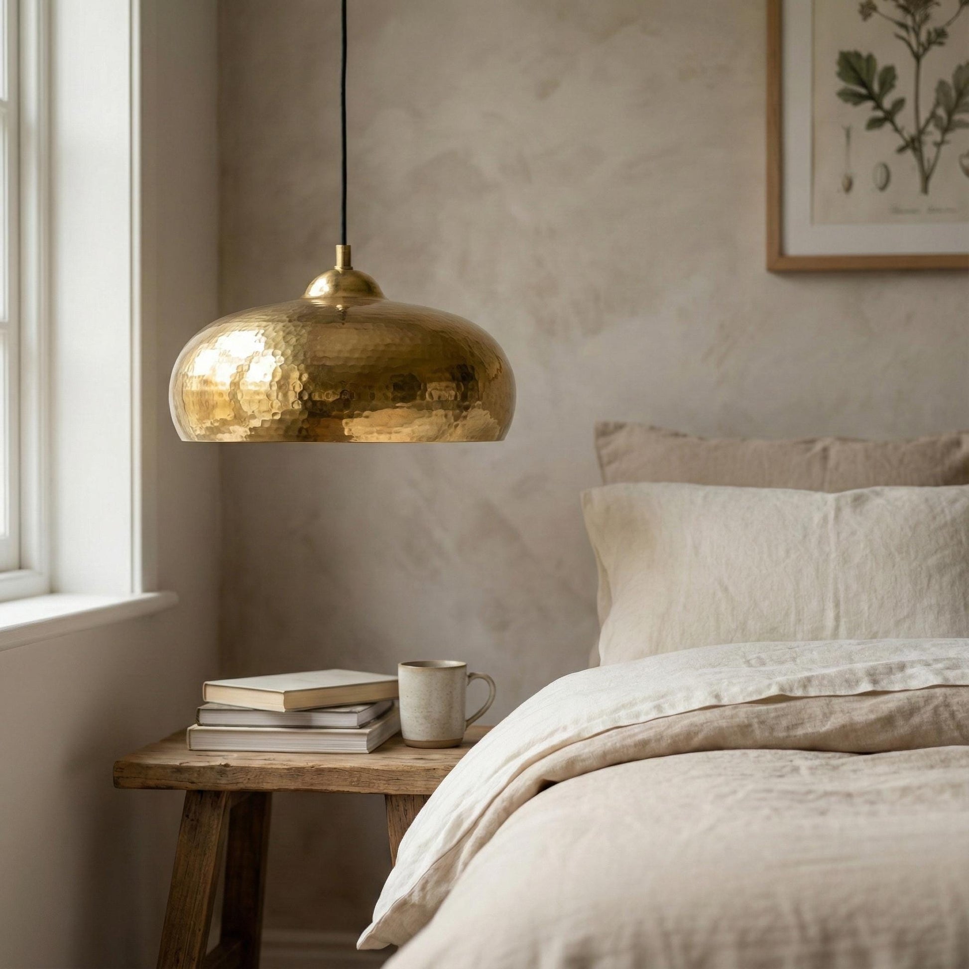 Neatly made bed with beige bedding, a wooden side table with books and a mug, and a gold pendant light in a bedroom.