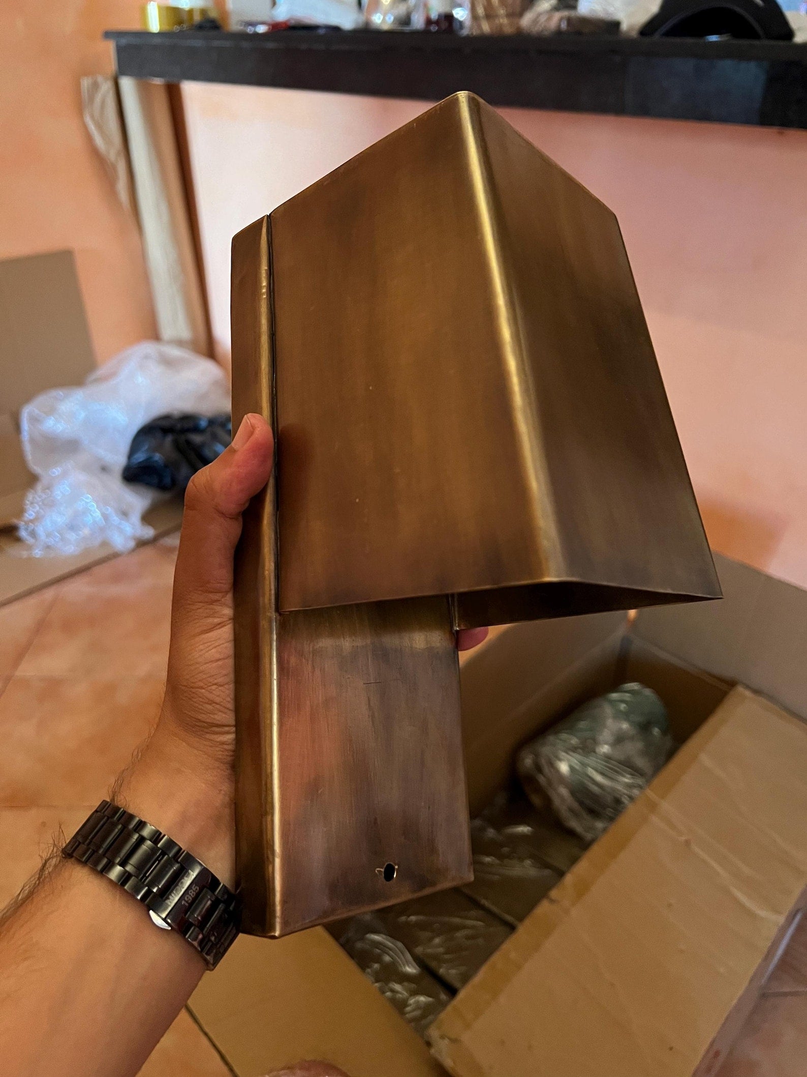 Hand holding a brass wall-mounted light fixture with a cardboard box and plastic bags in the background.