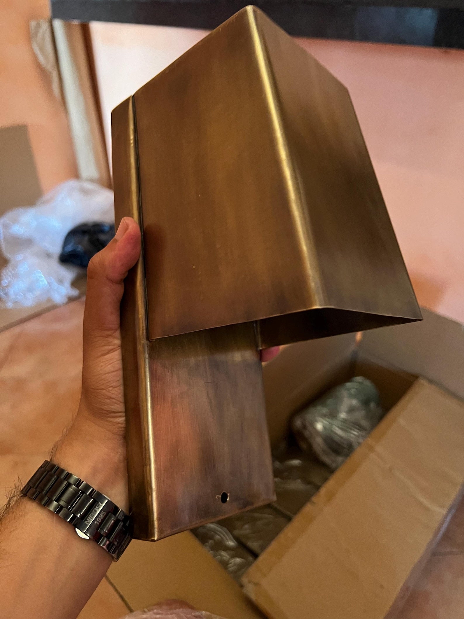 Bronze-colored metal bracket held by a hand with a watch, against a blurred indoor background.